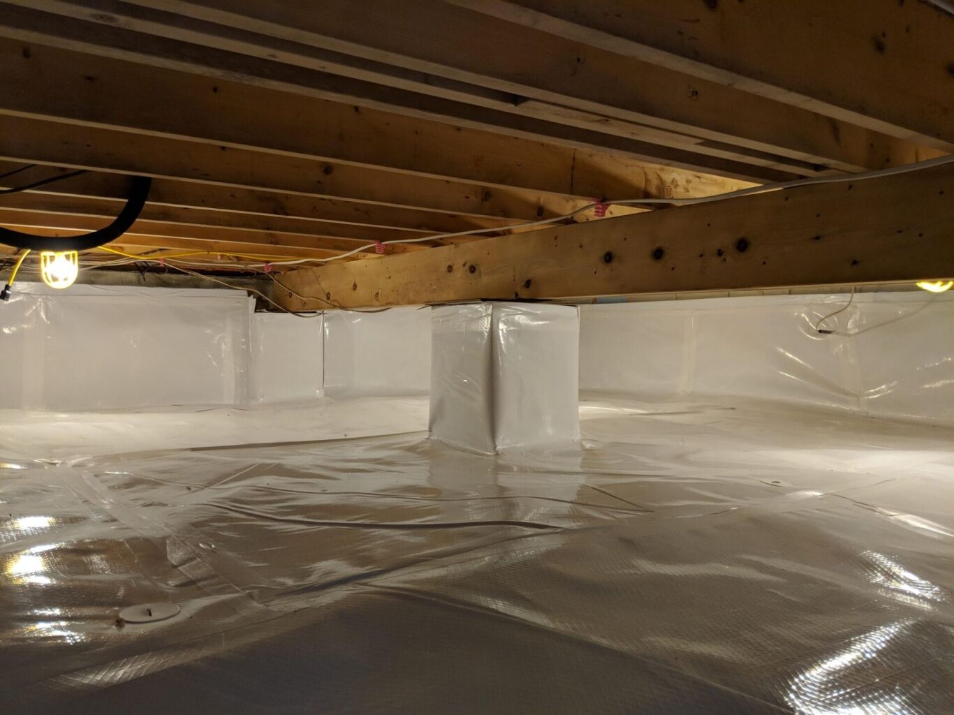 A clean, sealed crawl space with plastic vapor barrier covering the floor, walls, and support columns; exposed wooden beams above with utility lights.