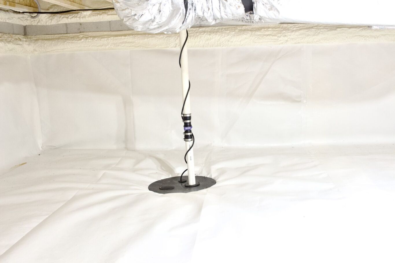 A sump pump discharge pipe extends through a circular opening in the white vapor barrier of a clean, encapsulated crawl space.
