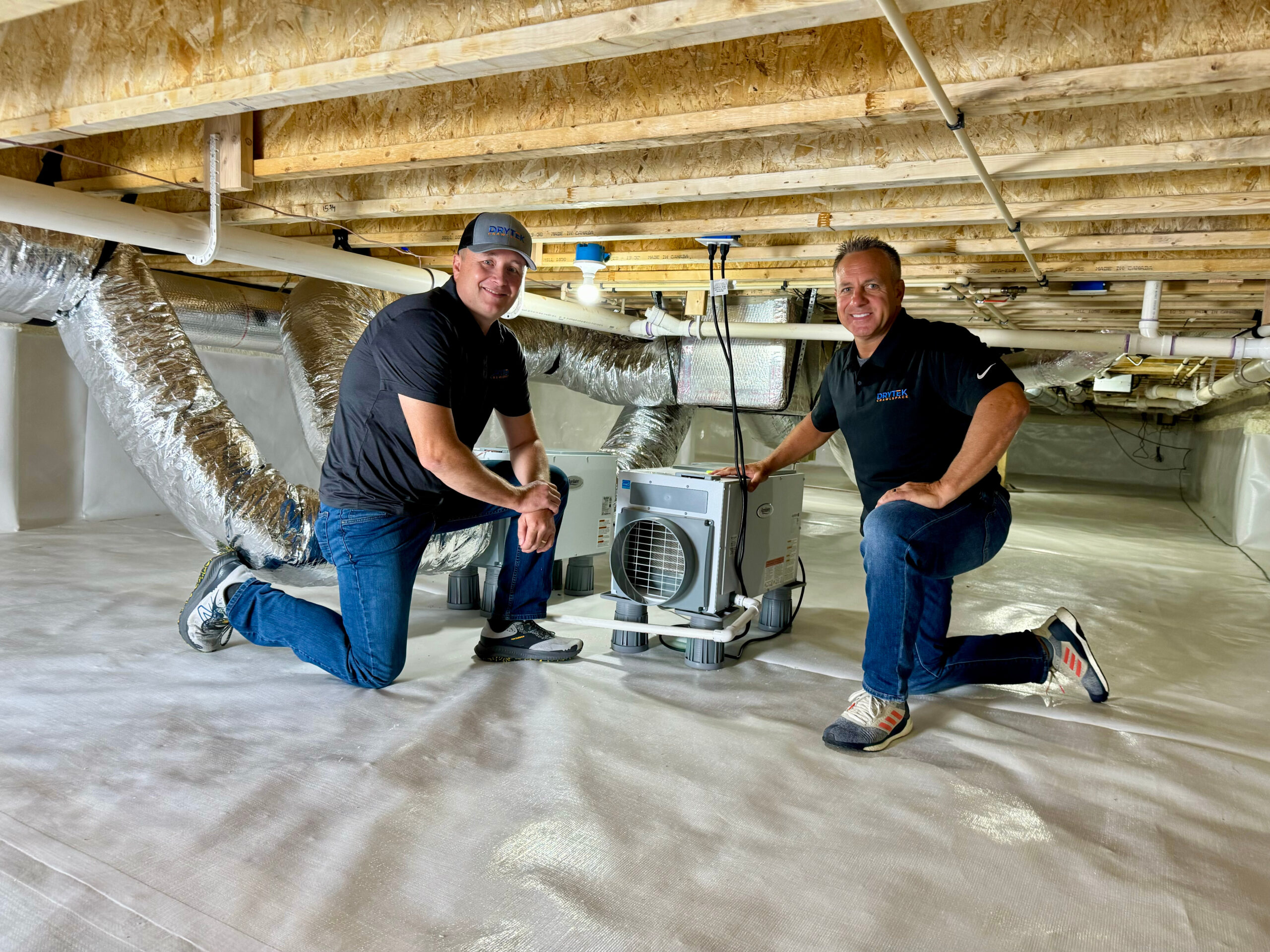 Two men kneel next to a dehumidifier in a clean, encapsulated crawl space with exposed wooden beams and insulated ducts overhead.