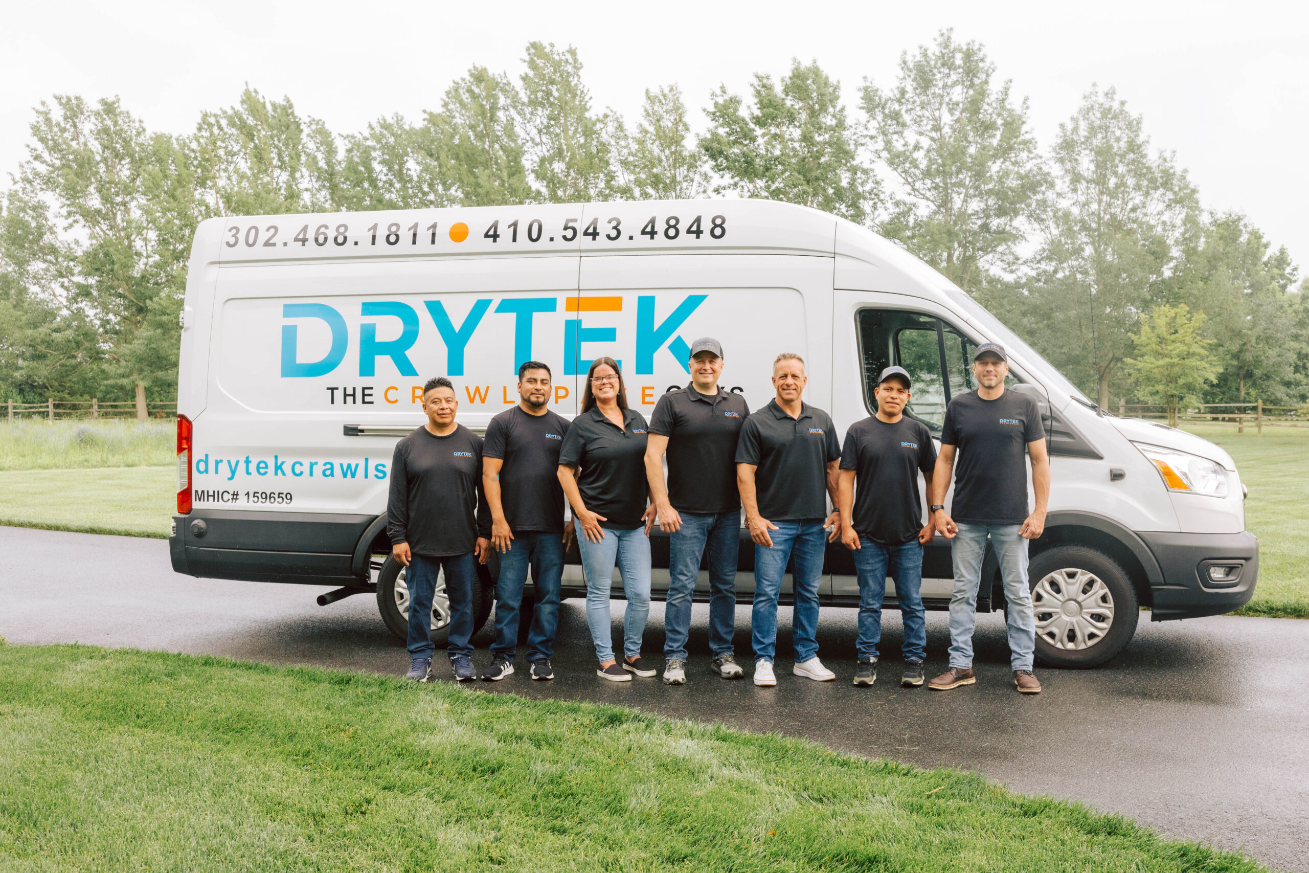 Eight people in black shirts and jeans stand in front of a white DryTek company van parked on a paved road with grass and trees in the background, representing our dedicated team—learn more on our About Us page.