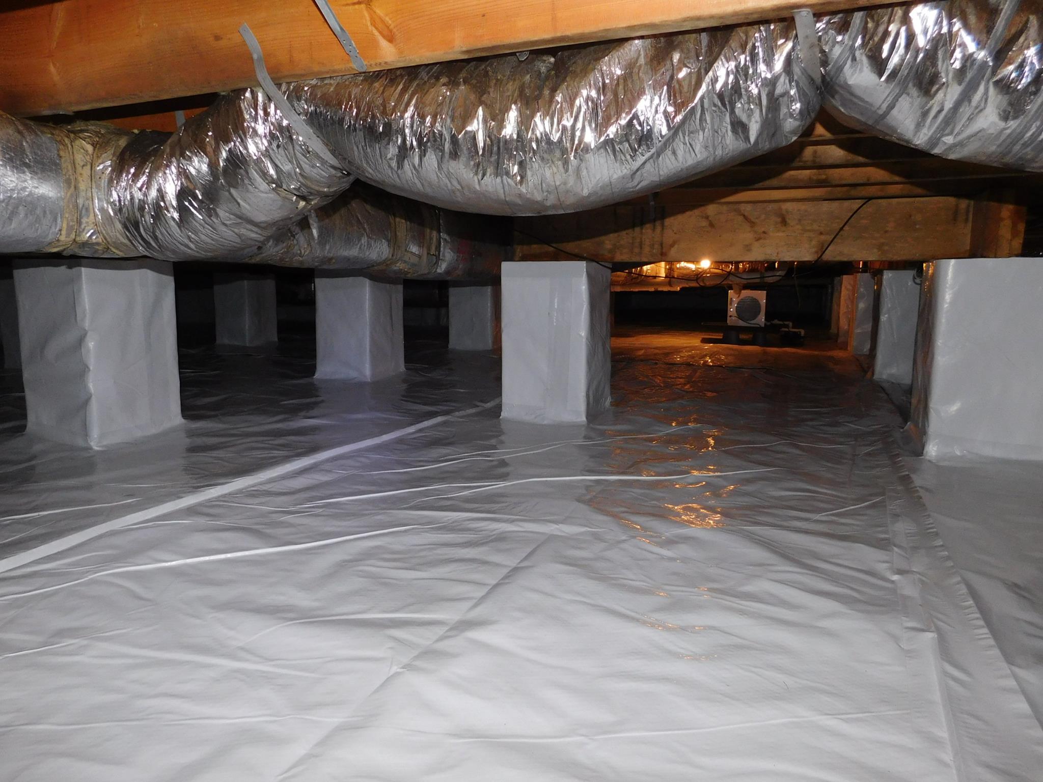A crawl space with ducts and wooden beams, with the floor and support pillars covered in a white vapor barrier plastic sheeting.