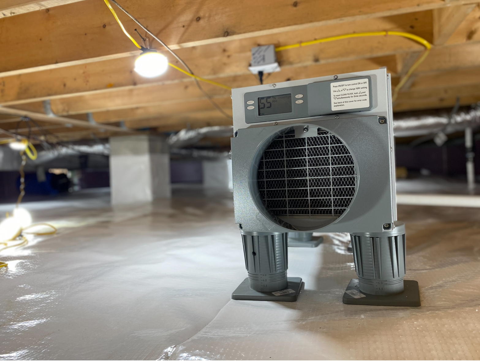A dehumidifier sits on a plastic-covered floor in a crawl space with exposed wooden beams and electrical wiring visible above.
