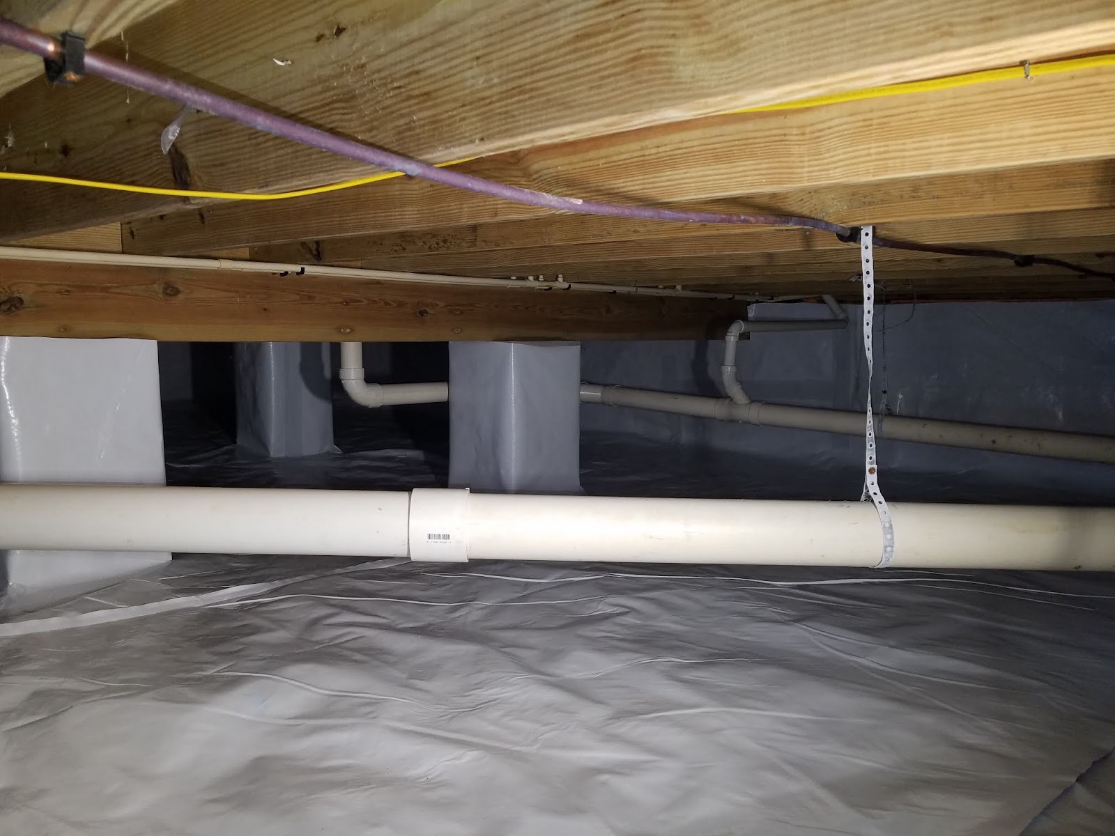 Crawl space with plastic vapor barrier lining the floor, exposed wooden beams above, and visible white PVC pipes and yellow electrical wiring.