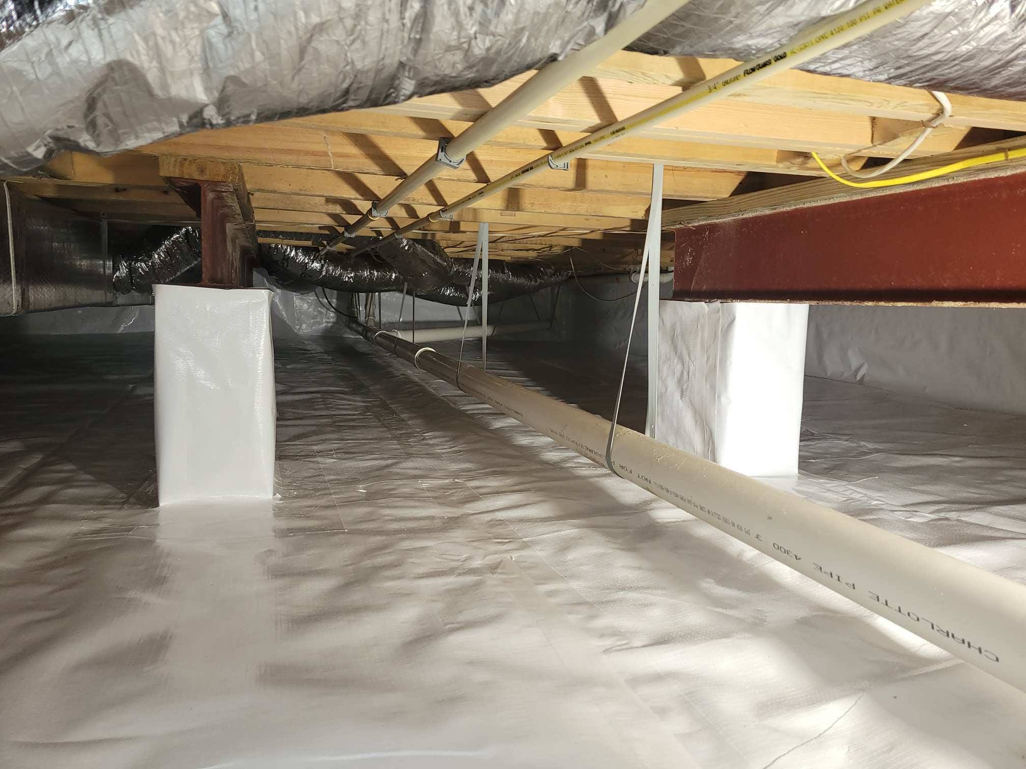 Crawl space with a plastic vapor barrier on the floor, exposed wooden beams, pipes, insulation, and electrical wiring overhead.