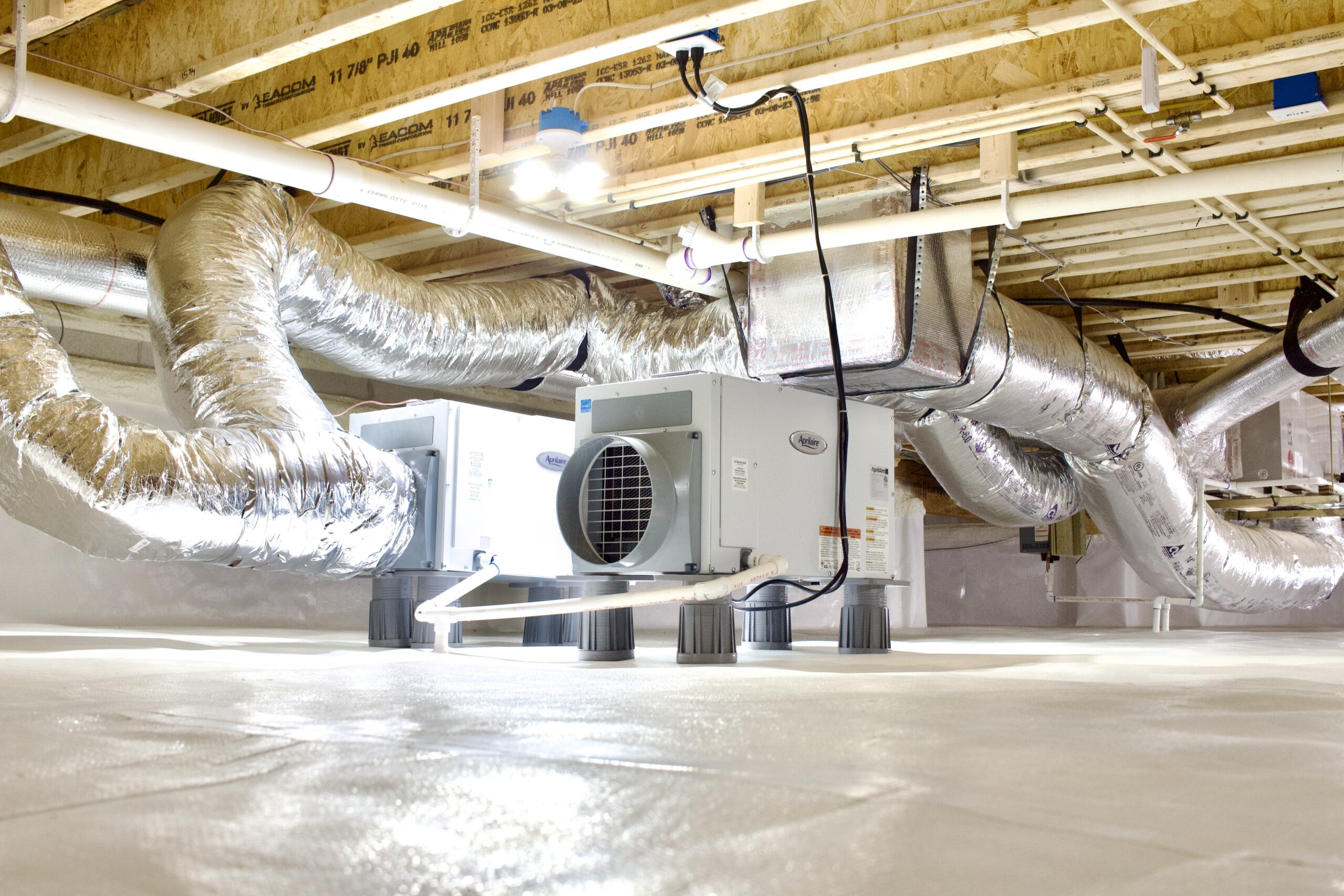 A clean, brightly lit crawl space with insulated HVAC ductwork and two dehumidifiers installed on a white vapor barrier floor.