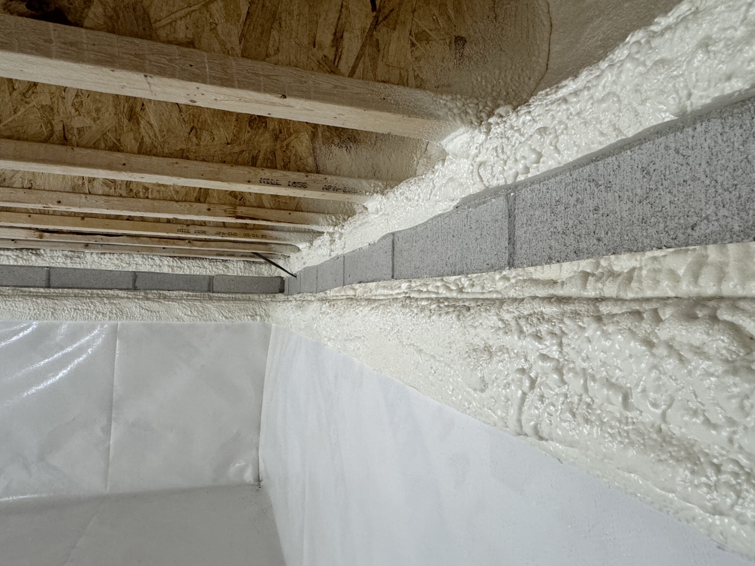 Wall section with spray foam insulation applied between wooden studs and concrete blocks, alongside a plastic vapor barrier and an unfinished wooden ceiling.