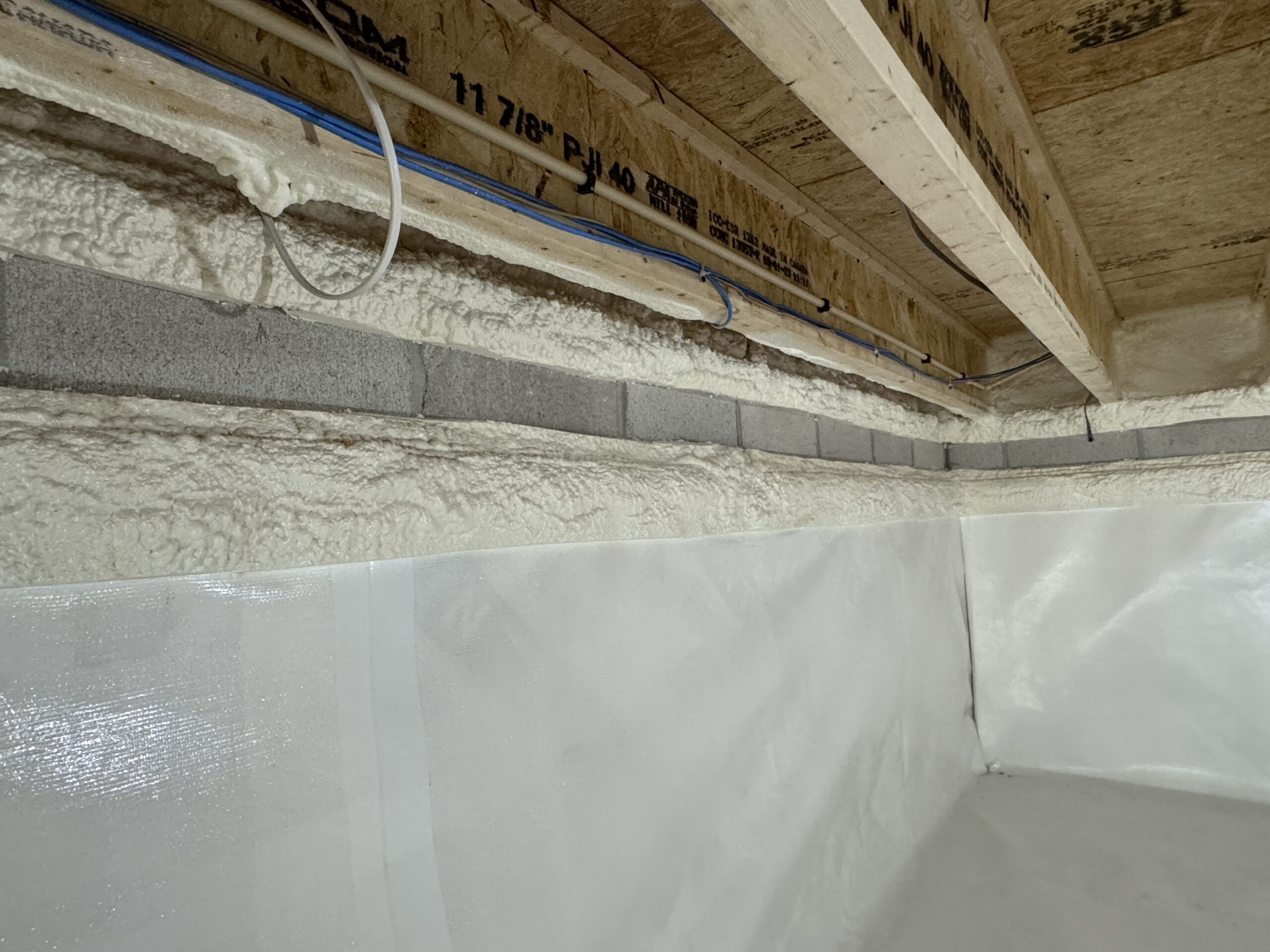 Insulated basement or crawl space wall with spray foam and vapor barrier, exposed ceiling joists, and visible electrical wiring.