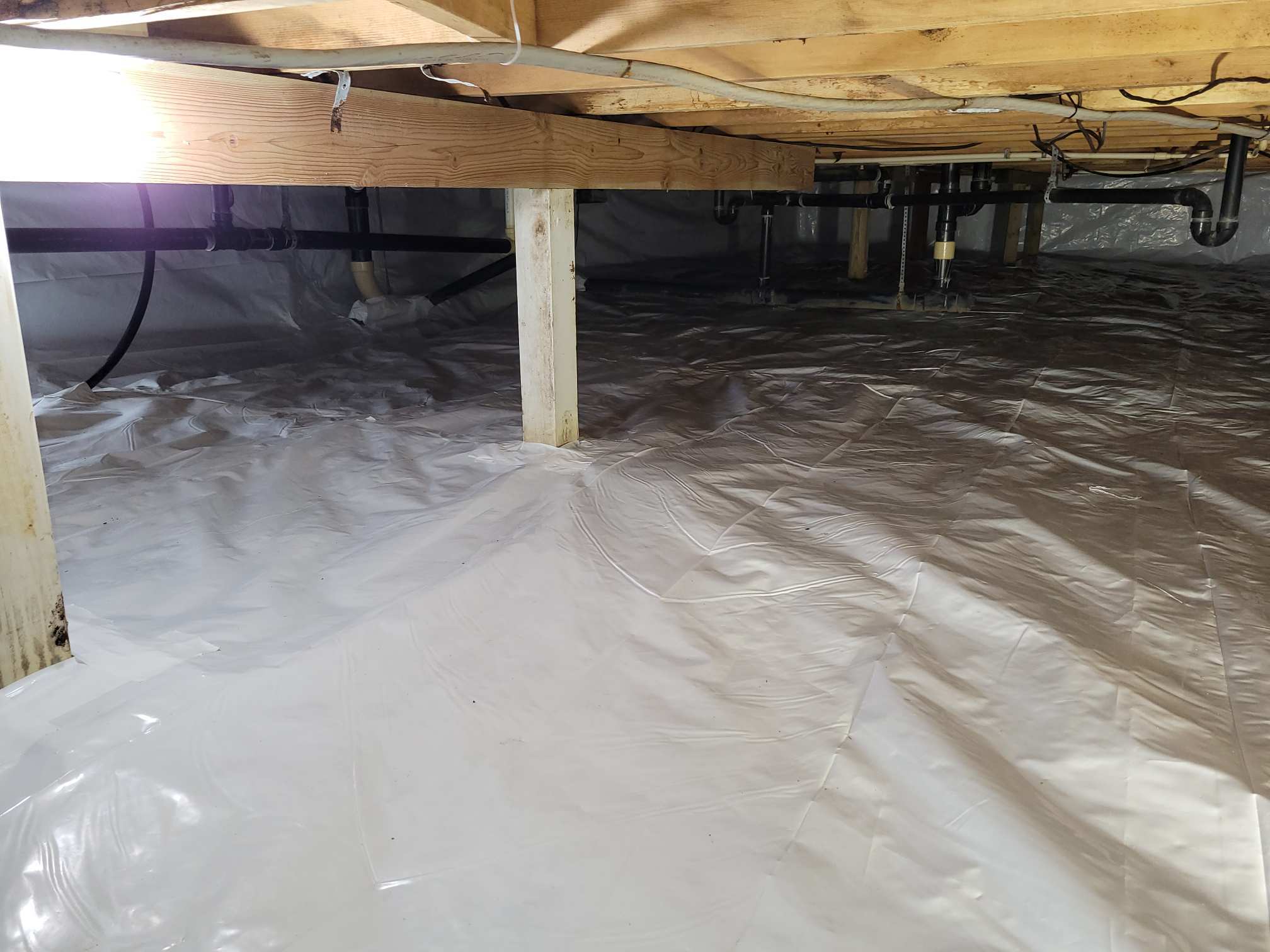 A crawl space under a house with wooden beams, support posts, and a white plastic vapor barrier covering the ground and walls.