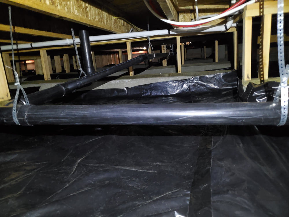 Black plastic pipes suspended under a wooden floor structure in a crawl space, with a plastic vapor barrier covering the ground.