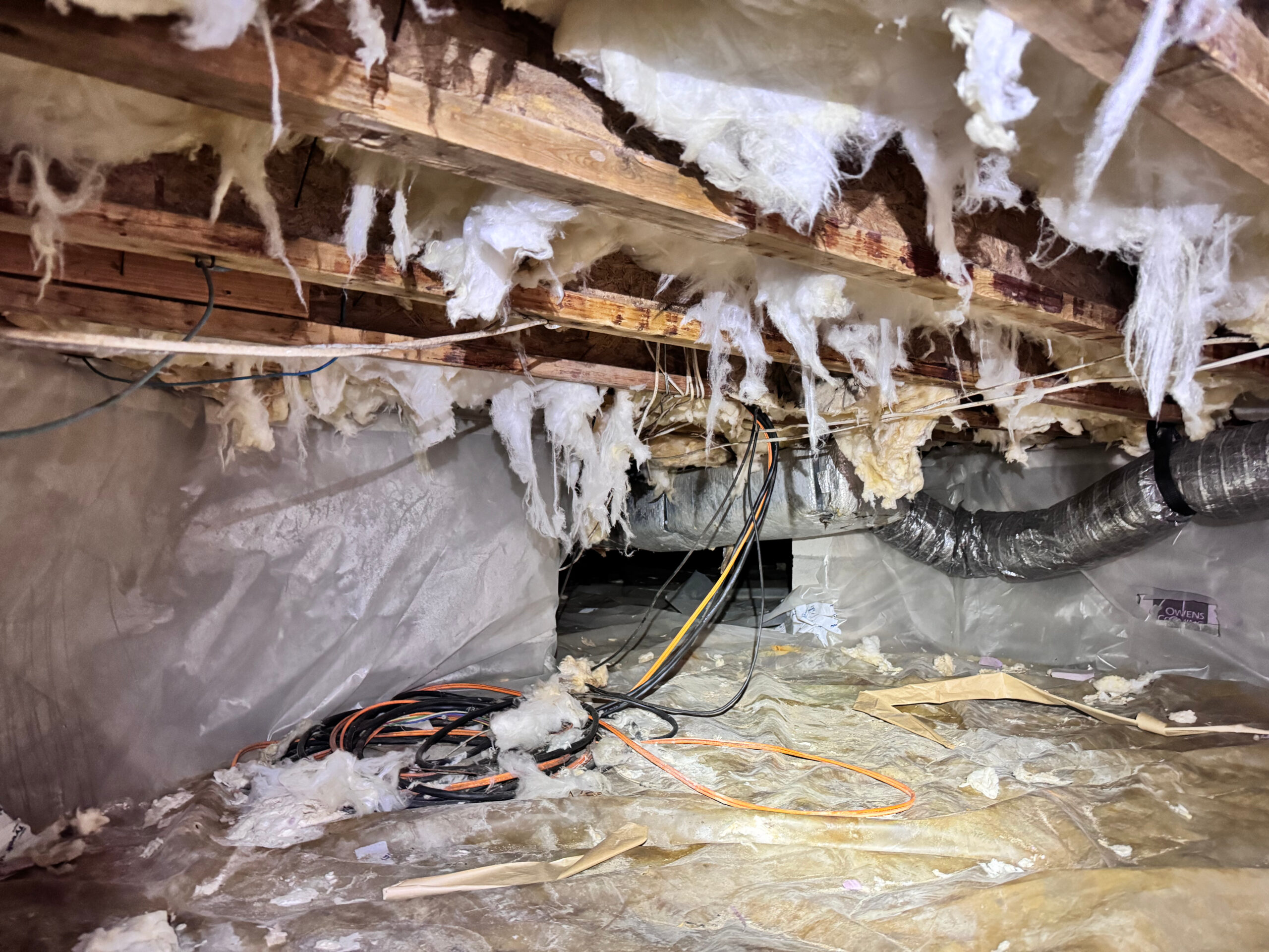 A crawl space with exposed insulation hanging from the ceiling, scattered debris, electrical cables, and ductwork.