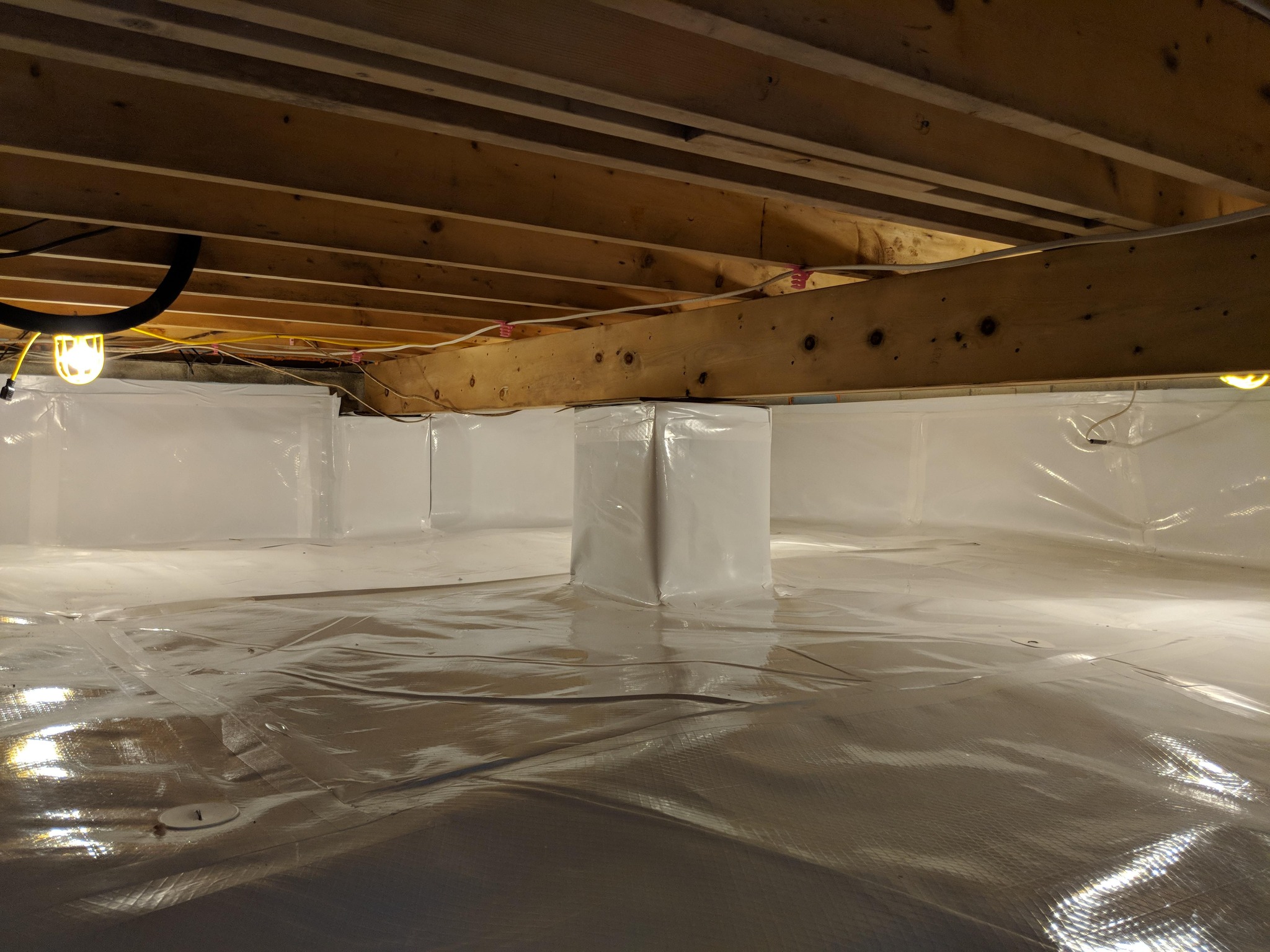 A clean, sealed crawl space with plastic vapor barrier covering the floor, walls, and support columns; exposed wooden beams above with utility lights.