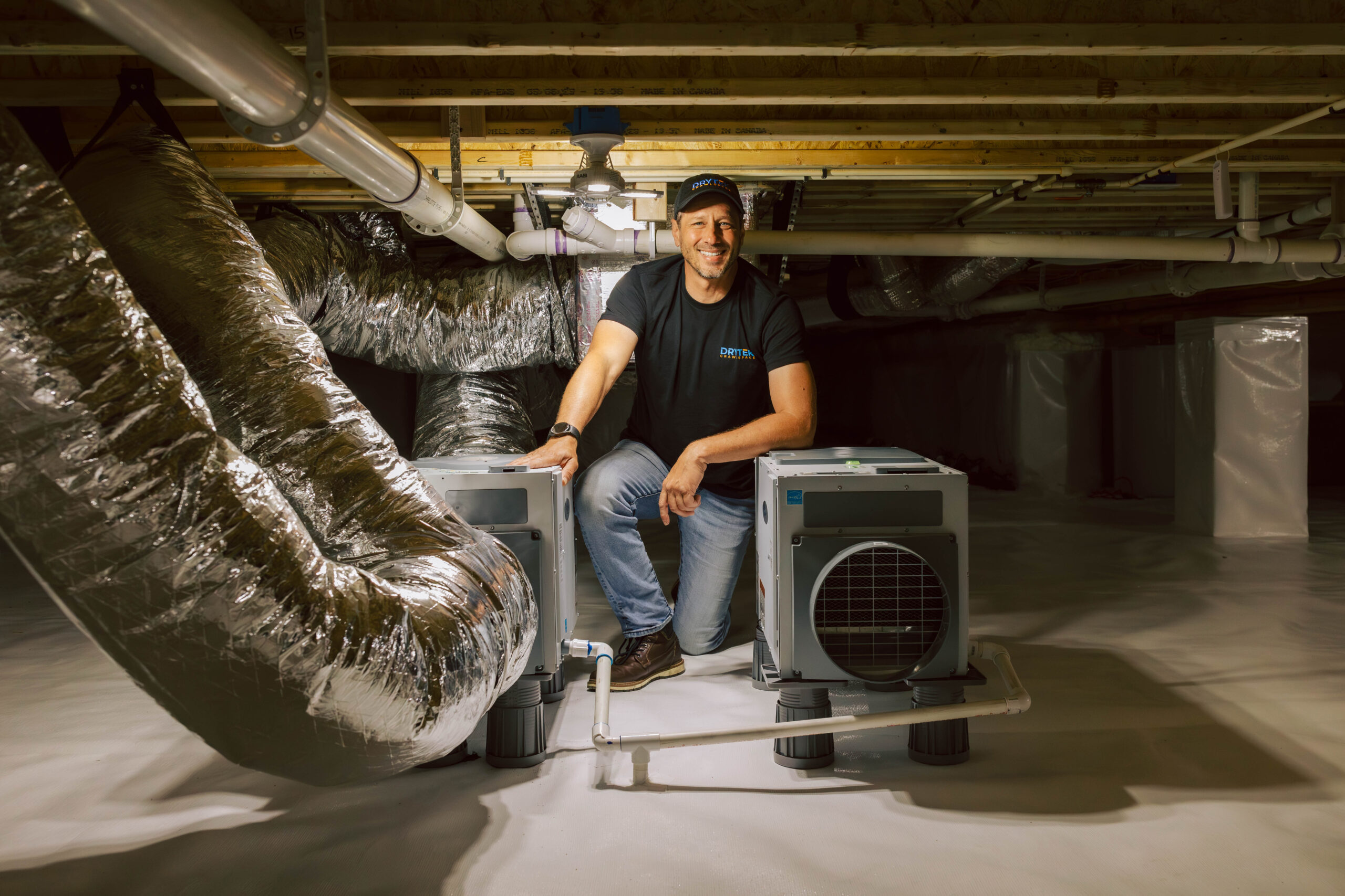 A person kneels in a clean, well-lit crawl space next to two air handling units and large insulated ventilation ducts, showcasing the professionalism featured in our About Us section.