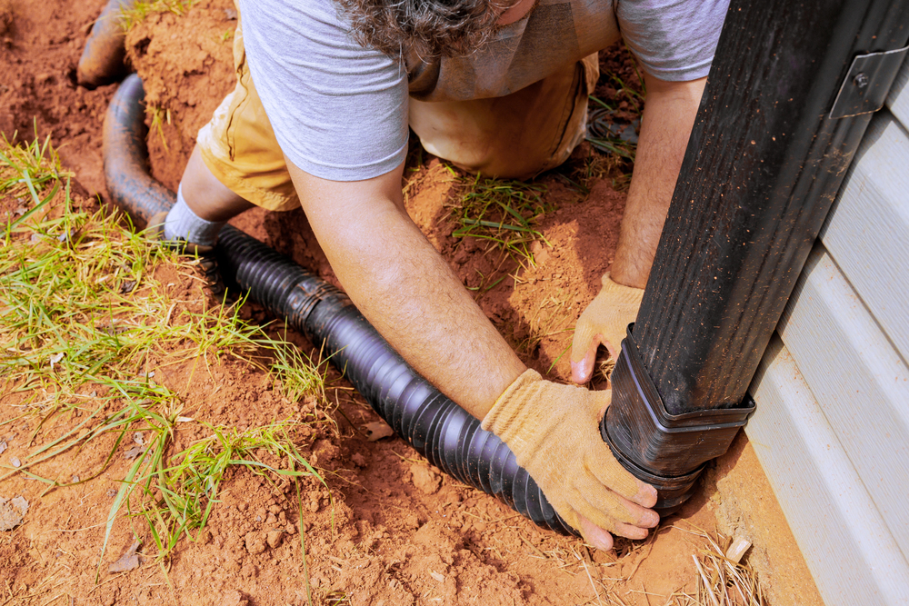 Person wearing gloves connects a black corrugated drainage pipe to a downspout next to a building, working in a dug-out area of soil and grass.