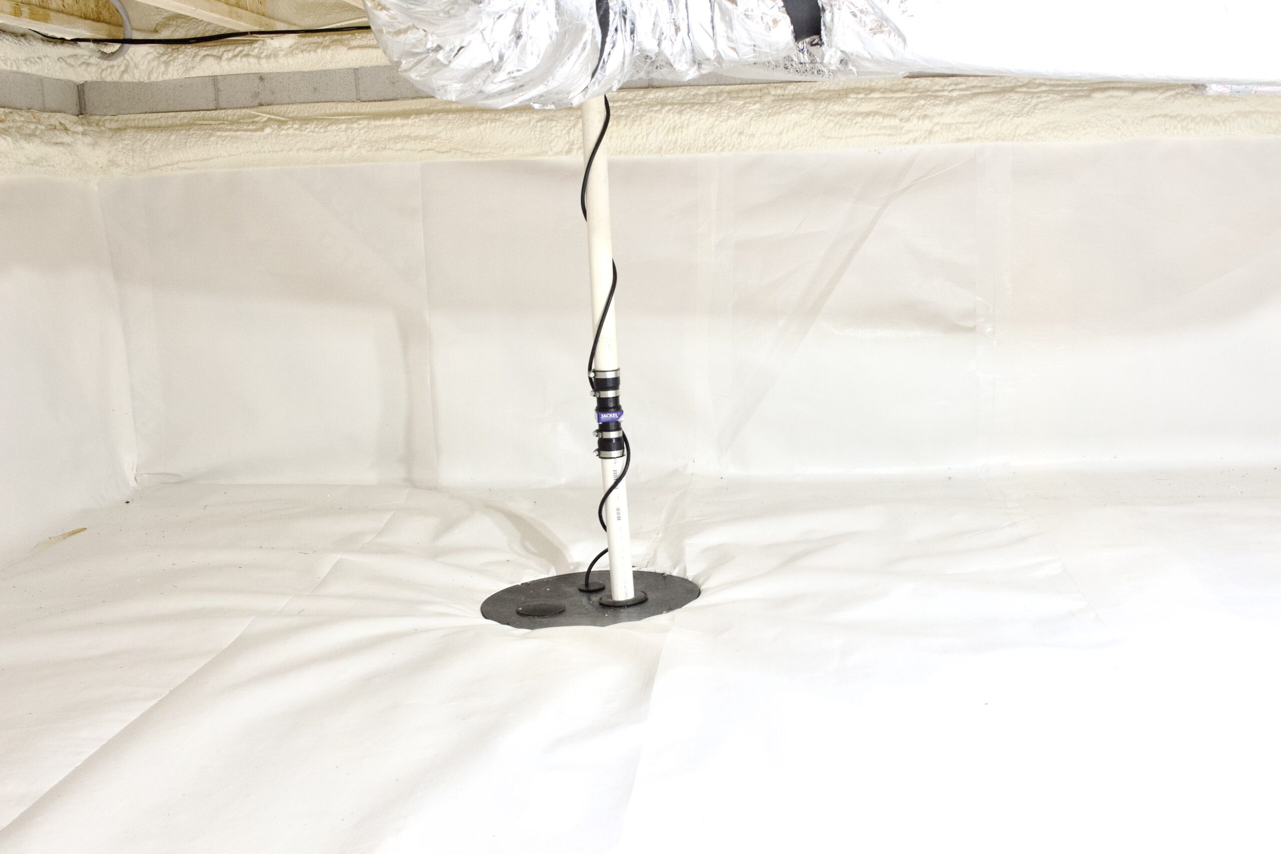 A sump pump discharge pipe extends through a circular opening in the white vapor barrier of a clean, encapsulated crawl space.